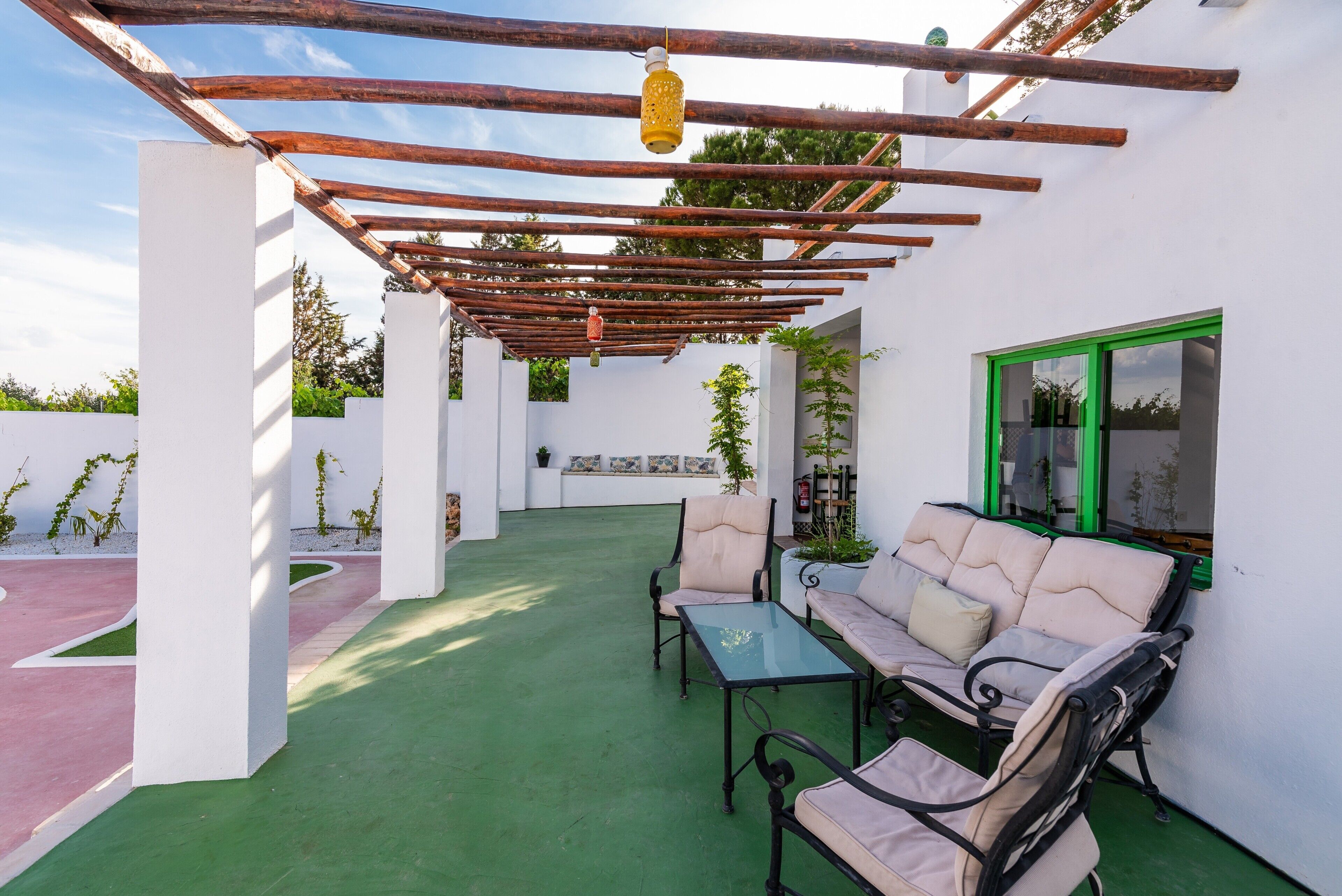 Un patio couvert avec une table et deux chaises, une lanterne suspendue et une fenêtre encadrée de vert.