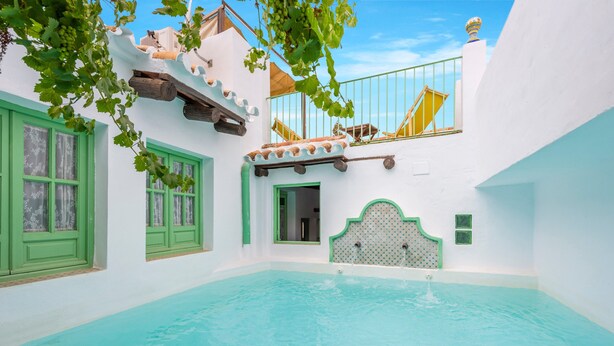 Zona de piscina con un edificio blanco, ventanas verdes y un balcón con barandillas amarillas.