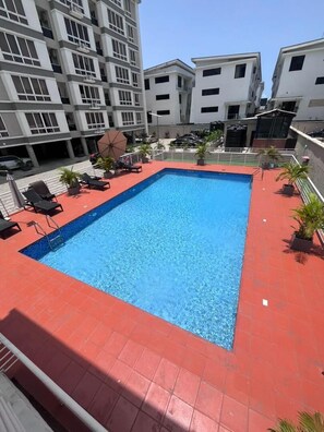 A rectangular pool with a red border, surrounded by lounge chairs and potted plants, with modern buildings in the background.