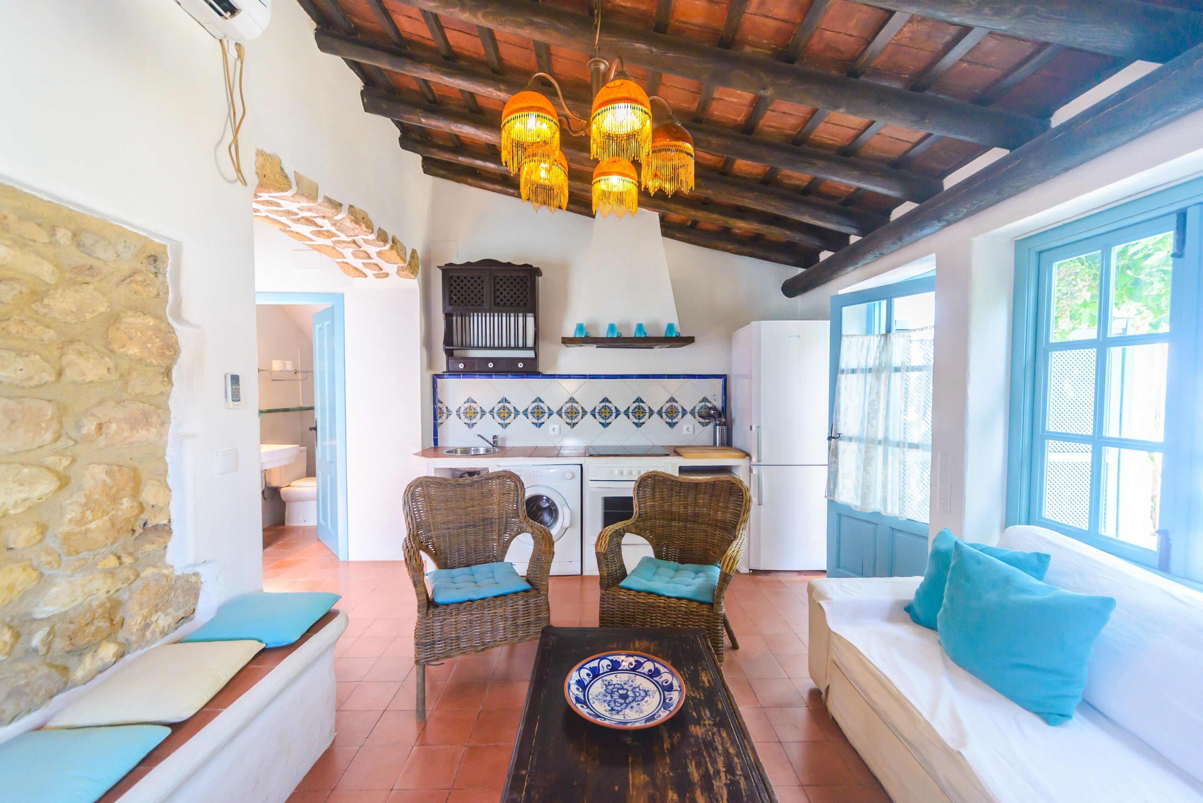 Un interior rústico con pared de piedra, muebles de mimbre, un sofá con cojines, una zona de cocina y techo de madera.