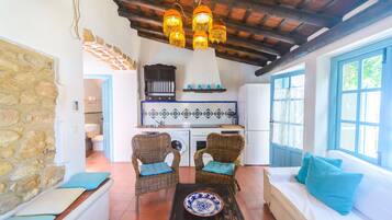 Un interior rústico con pared de piedra, muebles de mimbre, un sofá con cojines, una zona de cocina y techo de madera.