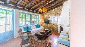 Una habitación acogedora con pared de piedra, un sofá blanco con cojines azules, sillas de mimbre, una mesa de centro de madera y una chimenea de piedra.