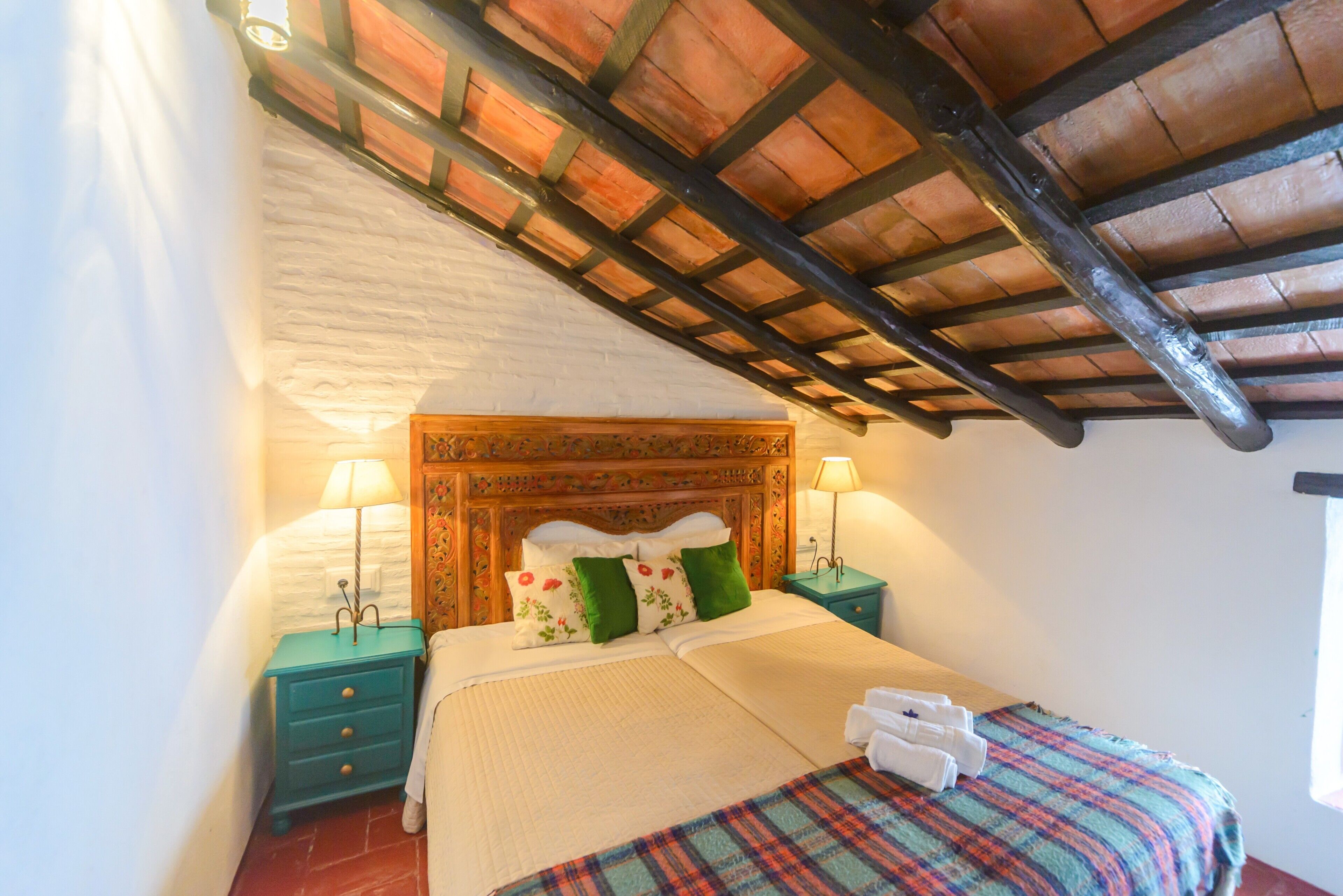 Une chambre à coucher avec une tête de lit en bois, deux tables de chevet avec des lampes, un lit recouvert d’une couverture à carreaux et un plafond carrelé.