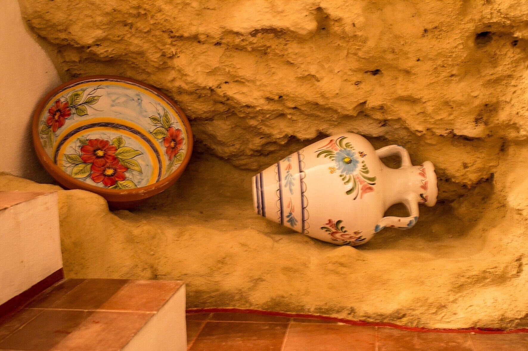 Una jarra y un cuenco de cerámica con motivos florales, ubicados en un hueco con paredes texturizadas.