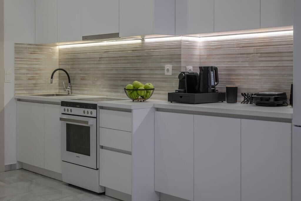 A modern kitchen with white cabinetry, a stainless steel range hood, a built-in oven, a coffee machine, and a bowl of green apples on the countertop.