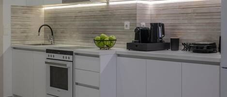 A modern kitchen with white cabinetry, a stainless steel range hood, a built-in oven, a coffee machine, and a bowl of green apples on the countertop.