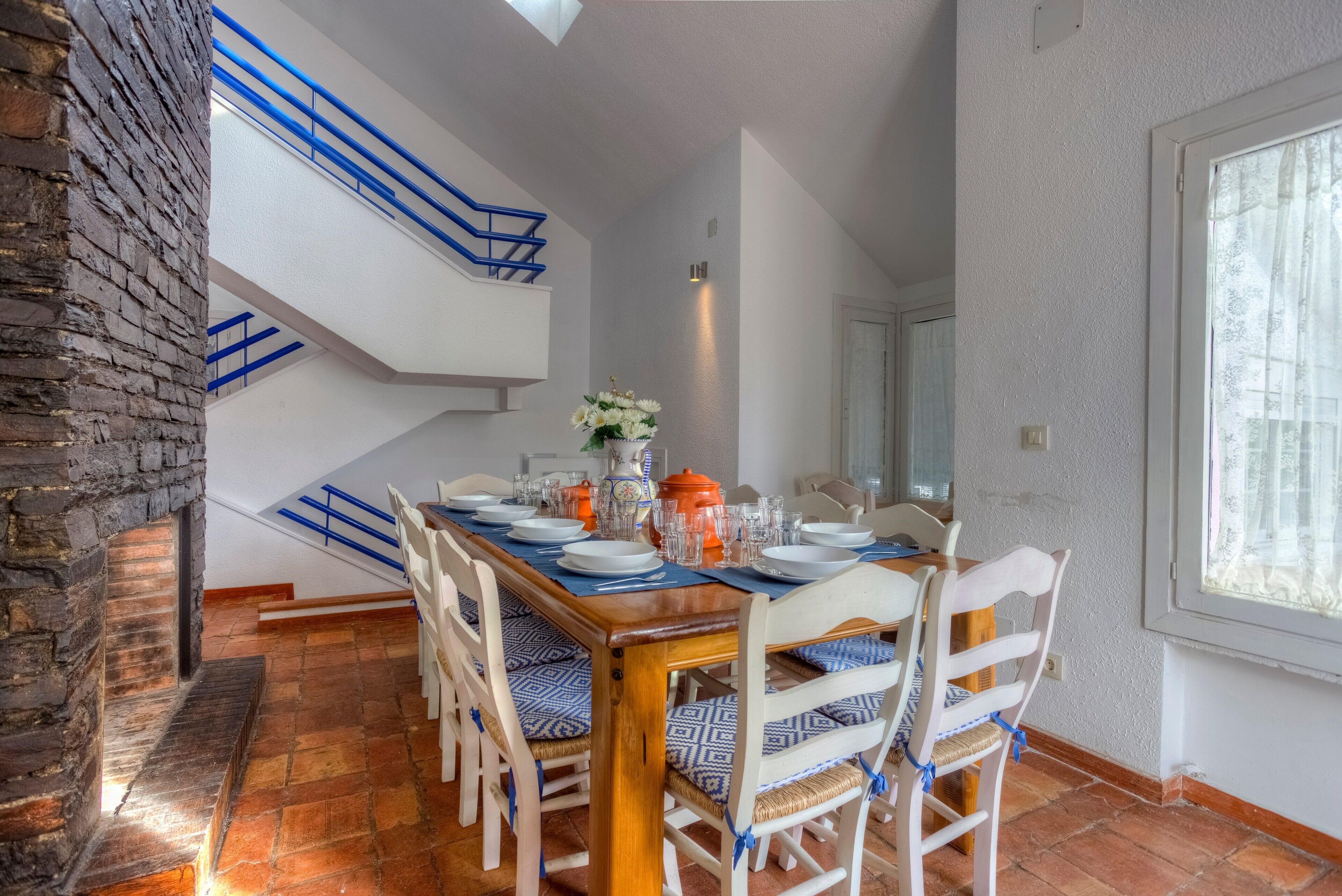 Un'area pranzo con un tavolo in legno apparecchiato per quattro persone, circondato da sedie bianche con cuscini blu. Si vedono anche un muro in mattoni e una scala con ringhiere blu.