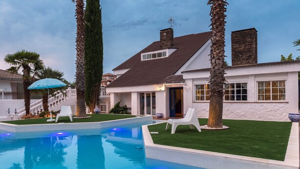 A swimming pool with white lounge chairs, palm trees, and a house with a stone facade.