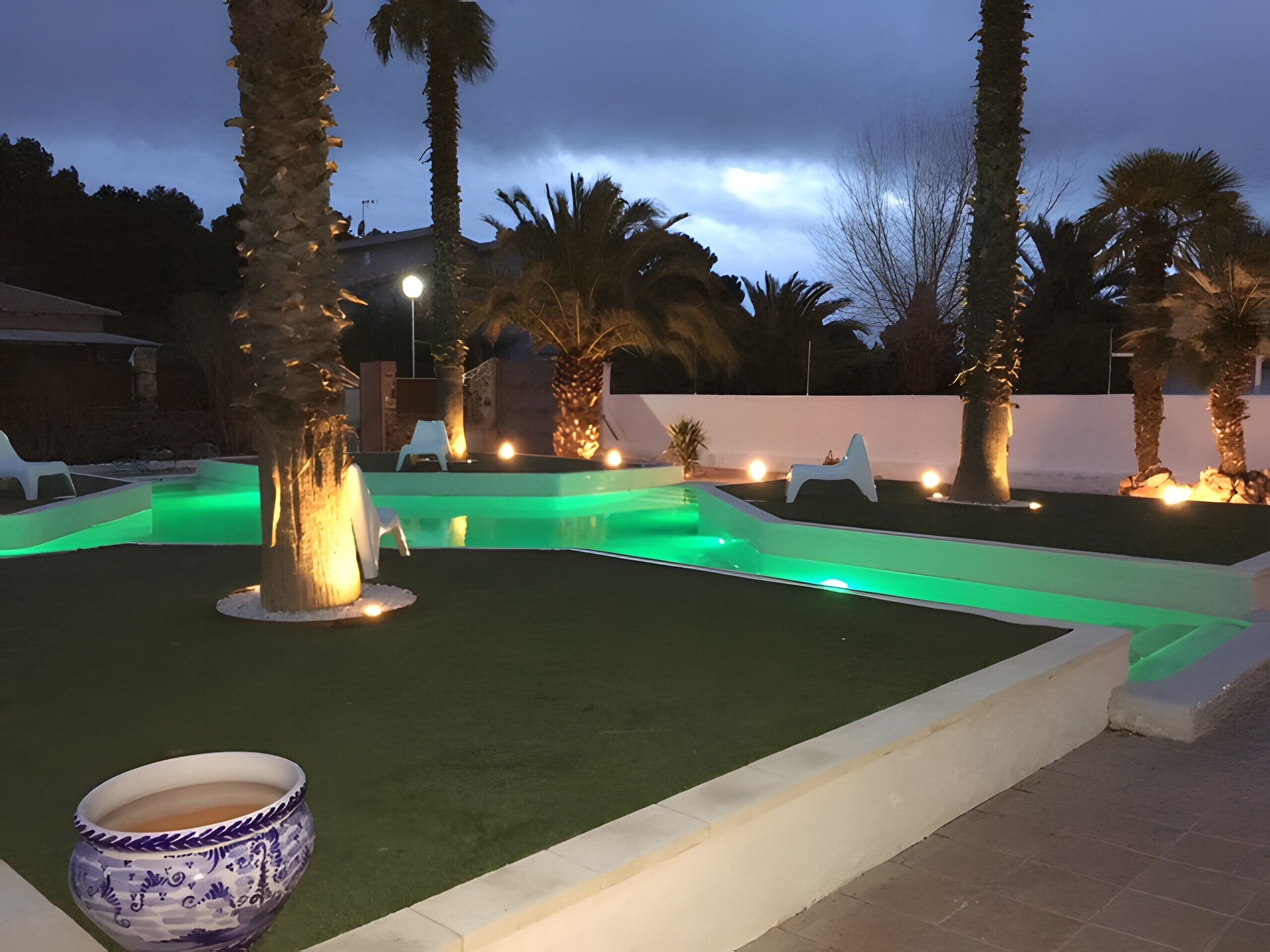 Une piscine éclairée de lumières vertes, avec des palmiers et un mur blanc en arrière-plan.