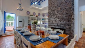 A dining area with a brick fireplace, a kitchen in the background, and a view of the outdoors.