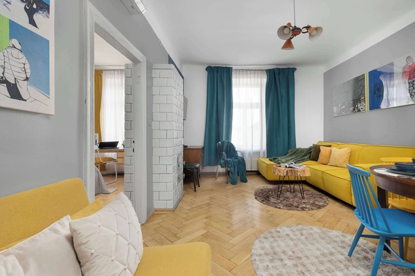 A spacious living room featuring a striking yellow corner sofa set against a light gray wall adorned with modern artwork. In the background, you can see the passageway to the bedroom and a table with blue chairs.