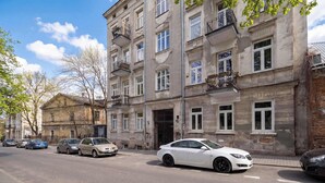 Un edifici de diverses plantes amb una entrada central, cotxes aparcats i un arbre de fulles verdes.