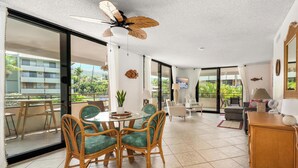 Un vaste espace de vie comprenant une table à manger, des chaises, un ventilateur de plafond et une vue sur un immeuble et la verdure environnante.