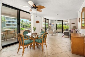 A spacious living area with a dining table, chairs, a ceiling fan, and a view of a building and greenery outside. - Summer Deal Beachfront Kona Condo with White Sand Beach Access (Kona)