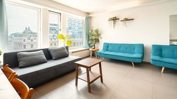 Un salón moderno con un sofá gris, un sofá azul, una mesita de madera y vistas a edificios a través de amplias ventanas.