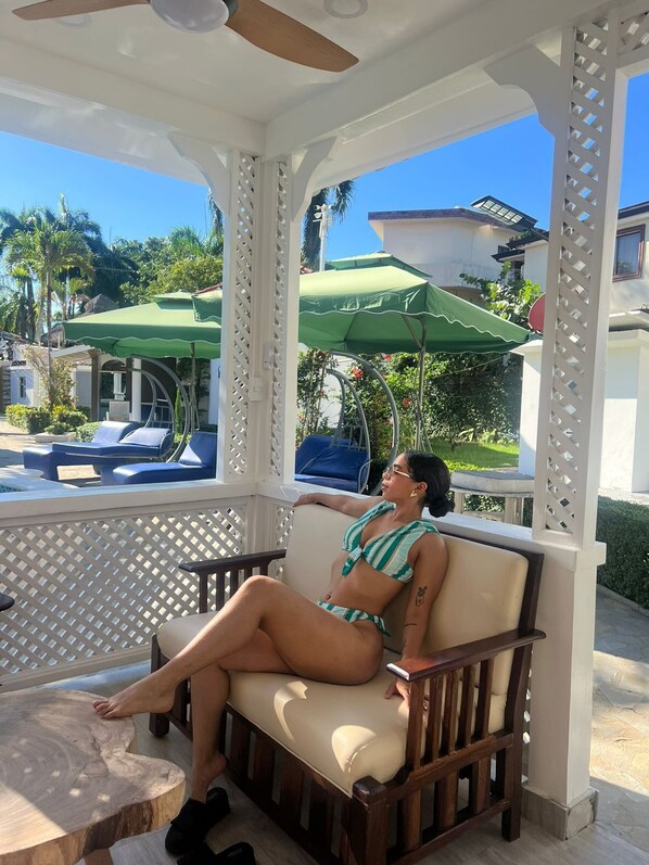 Uma pessoa relaxando em um banco de madeira, com vista para uma piscina e guarda-sóis em uma área externa coberta.
