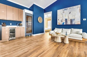 Un salon moderne avec un mur d’accent bleu, un canapé blanc, une table basse et un coin cuisine doté de meubles en bois et d’un réfrigérateur.