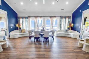 A spacious living room with a curved white sofa, a dining table with chairs, and a view through large windows with curtains.