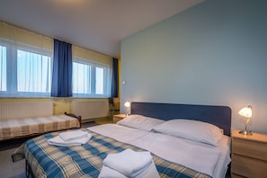 Un dormitorio con una cama grande, otra más pequeña, dos mesitas de noche con lámparas, una ventana con cortinas y un radiador.