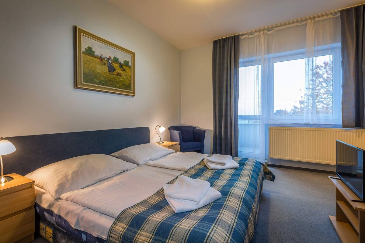 Habitación de hotel con dos camas, un escritorio con lámpara, una silla, televisión y un cuadro en la pared.