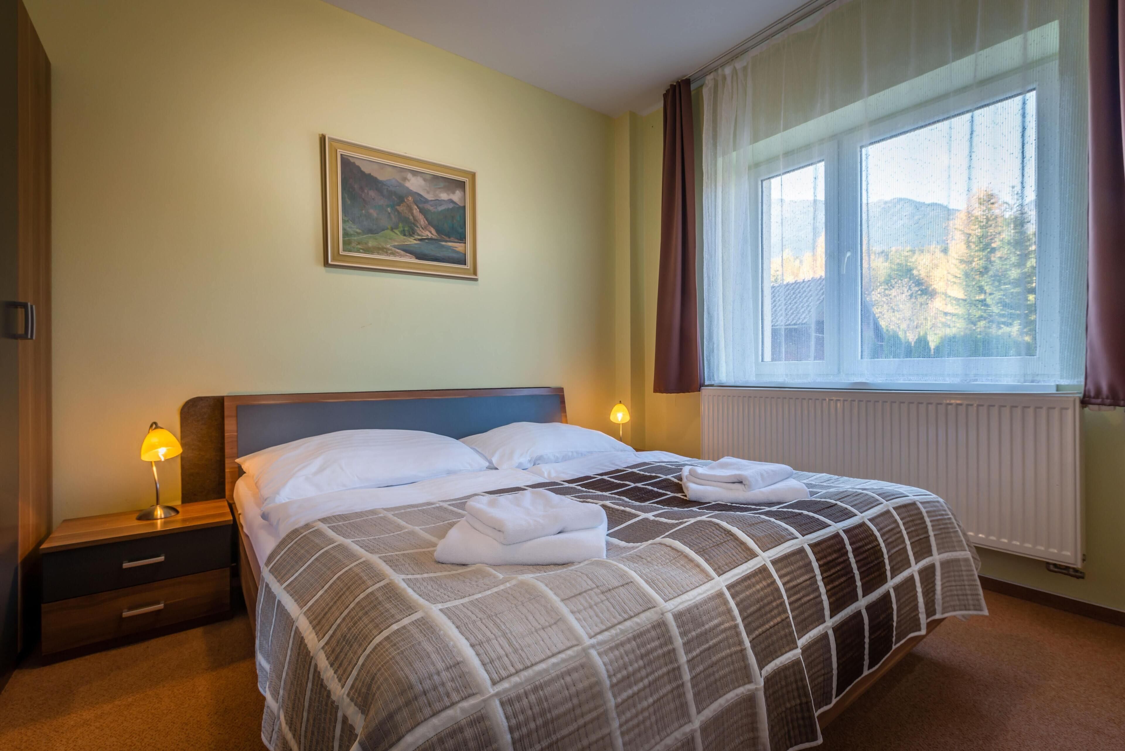 Een net opgemaakt bed met een geruit beddegoed, een bedlampje, een nachtkastje met lade, een ingelijste foto aan de muur en een raam met gordijnen.