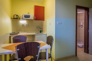 Una cocina compacta con microondas, tetera y armarios, una pequeña mesa de comedor con sillas, y una puerta de baño visible.