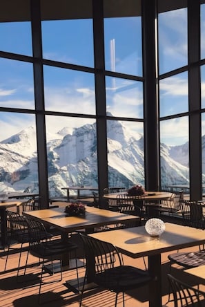 Ein Restaurant mit großen Fenstern und Blick auf die Berge. Tische mit Stühlen sind angerichtet.