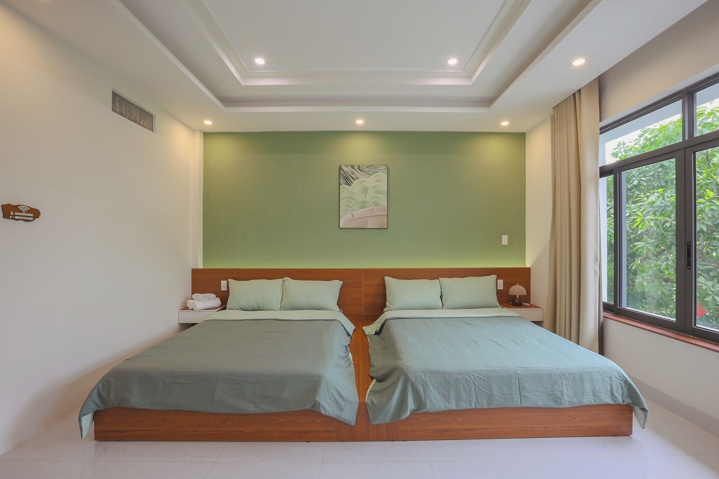 A double bed with a wooden headboard, a bedside table with a lamp, and a window with a view of greenery.