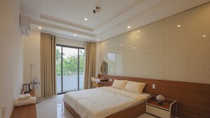 A modern hotel room with a large bed, a bedside table with a lamp, a desk with a fan, and a view of greenery outside. - TRÀ HOMESTAY CS3 (Hue)