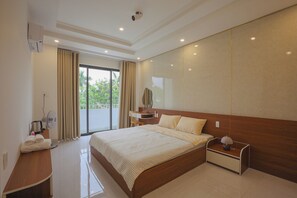 A modern hotel room with a large bed, a bedside table with a lamp, a desk with a fan, and a view of greenery outside. - TRÀ HOMESTAY CS3 (Phuong Vy Da)