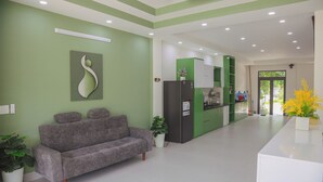 A reception area with a grey sofa, a white reception desk, green walls, and a ceiling with recessed lighting. - TRÀ HOMESTAY CS3 (Hue)