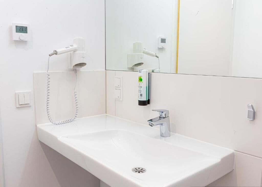 Un baño blanco con lavamanos, espejo, secador de pelo y dispensador de jabón.