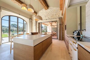 A modern kitchen with a large island, wooden beams, stone walls, and large windows. - Villa Lovea, charm from Provence (Saint-Rémy-de-Provence)