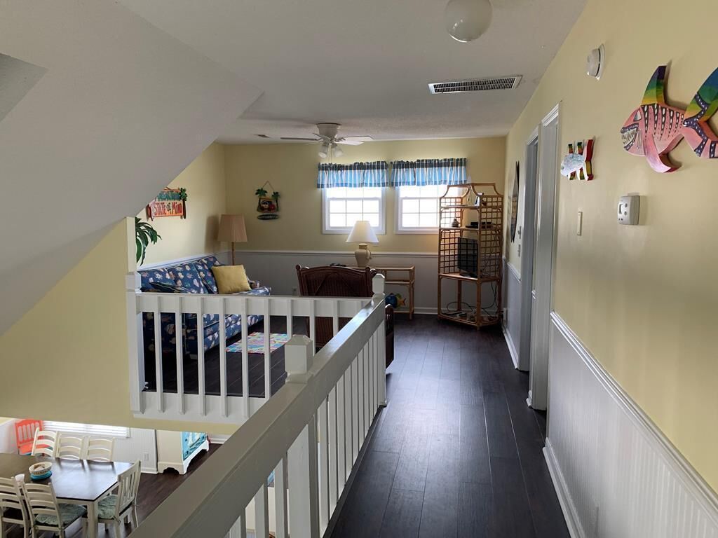 A room with a crib, a dining table, chairs, a window with curtains, a ceiling fan, and a wall-mounted shelf with decorative items.