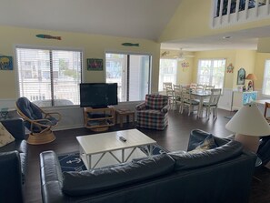 A living room with a sofa, a TV, a dining table with chairs, and a ceiling fan. - Canal Front Home - Sleeps 17! By Williamson Realty (Ocean Isle Beach)