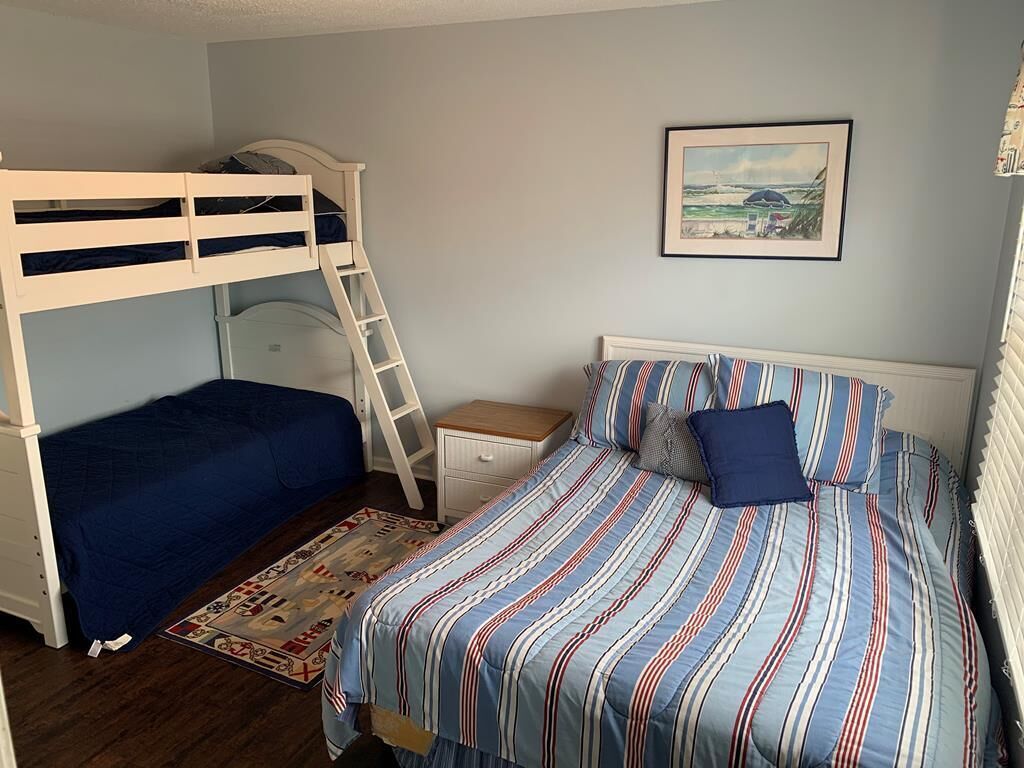 Une chambre avec des lits superposés, un lit simple avec une literie à rayures, une petite table de chevet et un tableau encadré au mur.
