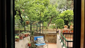 Een balkon met een stoel, plantenpotten en uitzicht op een tuin.