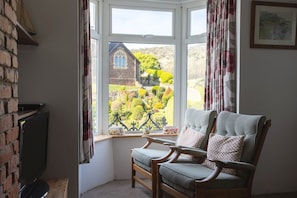 A room with a large bay window, a wooden chair, a television, and a brick wall.