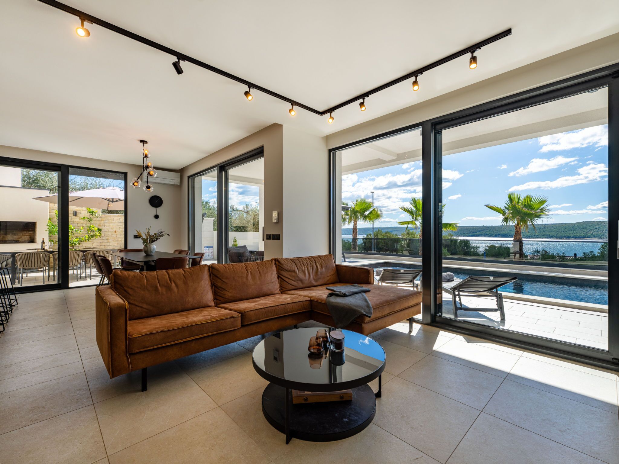 Uma sala de estar moderna com um sofá de couro, uma mesa de centro redonda e grandes janelas com vista para uma piscina e paisagem.