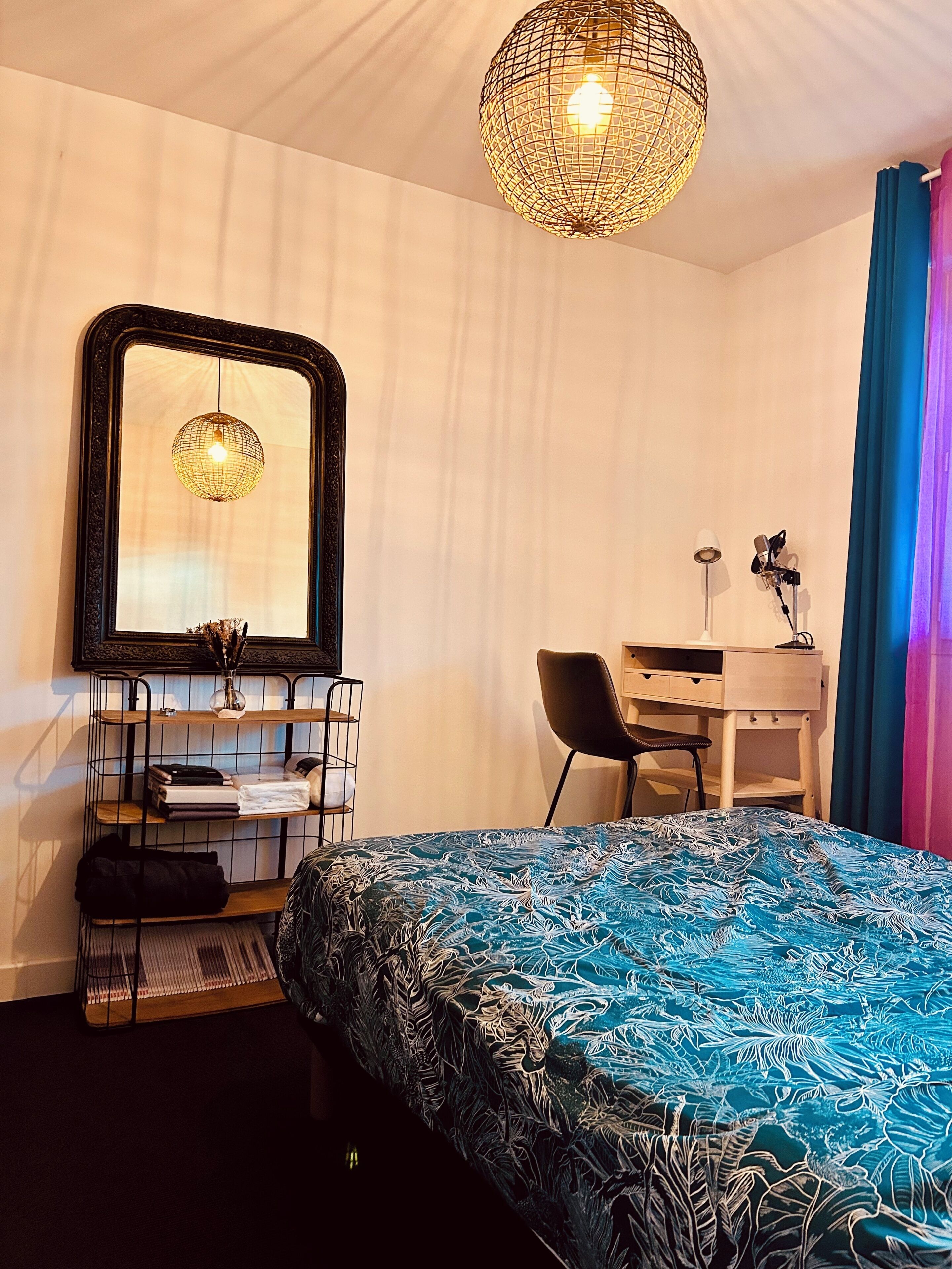 A bedroom with a bed, a desk, a chair, a mirror, and a hanging light fixture.