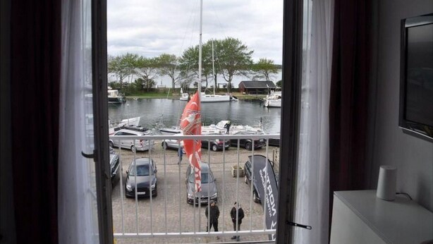 Vista da un balcone su una marina con barche, una bandiera e alcune auto.