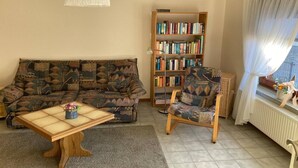 A living room with a patterned sofa, a wooden coffee table, a bookshelf, and a chair. - Ferienwohnung in Orth auf Fehmarn (Orth)