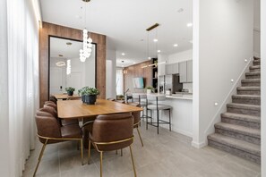 A modern kitchen with a central island, a dining area with a wooden table and leather chairs, and a staircase leading to an upper level. - 5BD Townhome: Prime Location, Game Room & Pool! (Orlando)