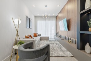 A modern living room with a sofa, a coffee table, a floor lamp, and a flat-screen TV mounted on the wall. - 5BD Townhome: Prime Location, Game Room & Pool! (Orlando)