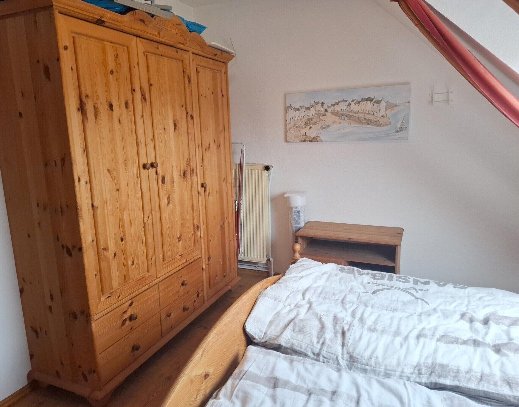 A bedroom with a wooden wardrobe, a bed with white bedding, a small wooden nightstand, a radiator, and a wall-mounted painting.