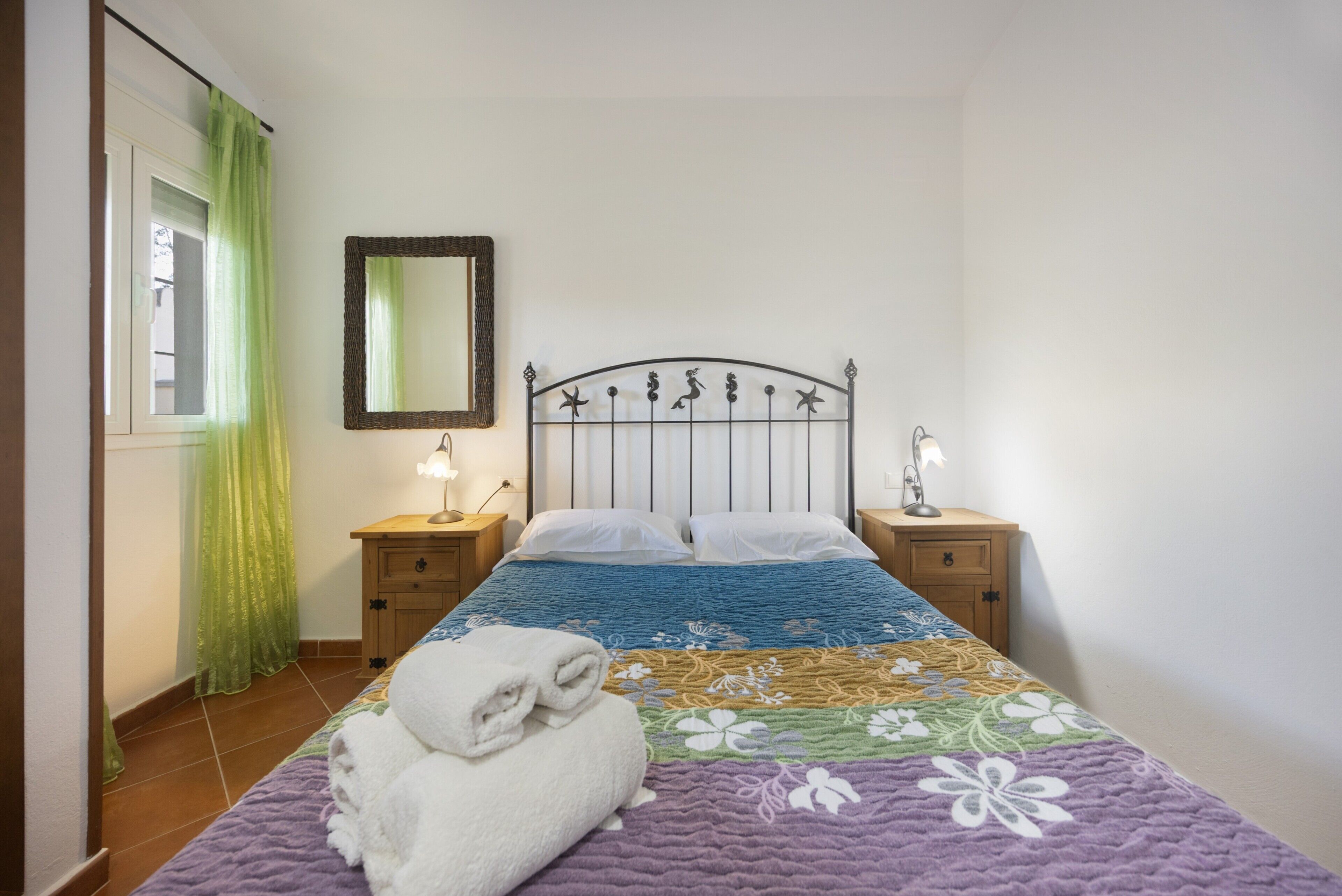 A bedroom with a bed, two bedside tables, a mirror, a window with curtains, and a towel on the bed.