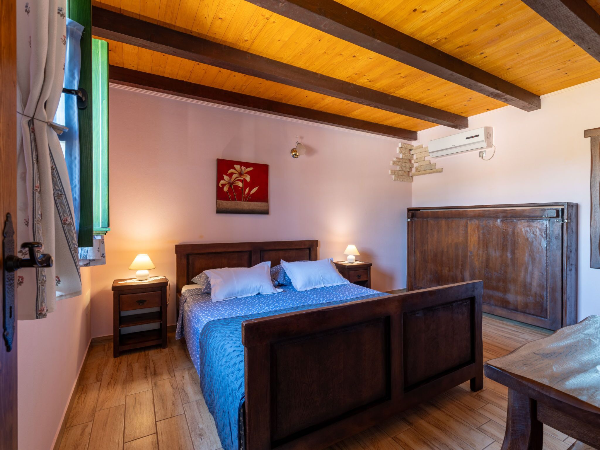 A bedroom with a wooden bed, two bedside tables, a nightstand, a wall-mounted air conditioner, and a large wooden wardrobe.