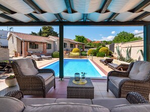 Ein überdachter Außenbereich mit Pool, Korbmöbeln und Blick auf benachbarte Häuser.