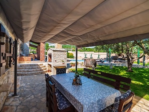 An outdoor patio with a table and benches, stone flooring, and a covered area with a view of a pool and greenery. - Stone villa Tea with pool by Interhome (Posedarje)