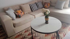 Um sofá bege com almofadas listradas e estampadas, uma mesa branca redonda com um vaso de flores e um tapete com padrão geométrico em zigue-zague.